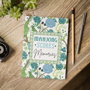 May include: A notebook with a floral design and the words "Mahjong Scores Memories" in a decorative frame. The cover features green birds, blue flowers, and branches. A pencil and pen rest on a wooden surface.