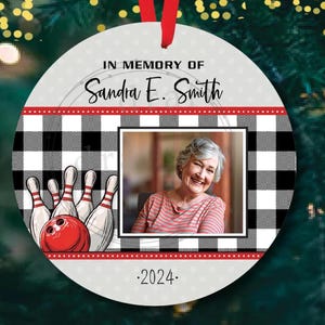 May include: A white round ornament with a black and white checkered pattern. The ornament has a red ribbon at the top and a photo of a woman in the center. The text "In Memory of Sandra E. Smith" is at the top of the ornament and the text ".2024." is at the bottom. The ornament also features a bowling pin and ball graphic.