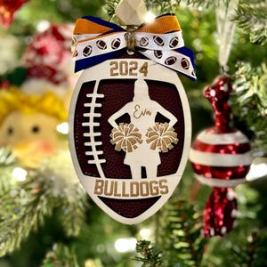 May include: A wooden football-shaped Christmas ornament with a maroon center and a cheerleader silhouette. The ornament has the text "2024", "Eva", and "Bulldogs". It is decorated with a blue, white, and orange ribbon and a wooden bead.
