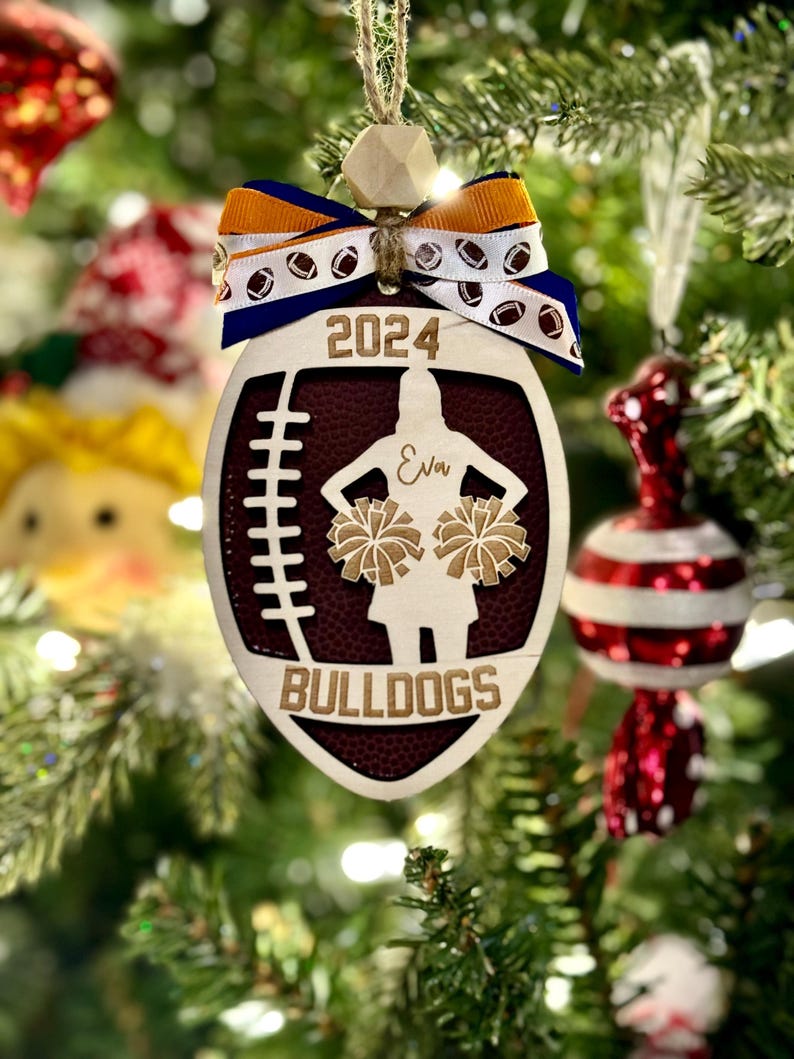 May include: A wooden football-shaped Christmas ornament with a maroon center and a cheerleader silhouette. The ornament has the text "2024", "Eva", and "Bulldogs". It is decorated with a blue, white, and orange ribbon and a wooden bead.