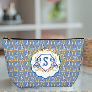May include: A blue cosmetic bag with a repeating pattern of dogs and gold rope. The bag features a central emblem with the letter "S" and two dog illustrations. The bag has a gold zipper.