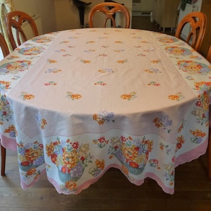 Può includere: Una tovaglia ovale con un motivo floreale. La tovaglia ha un centro a righe rosa e bianche, un bordo bianco con cesti floreali e un bordo smerlato rosa. Sedie in legno circondano il tavolo.