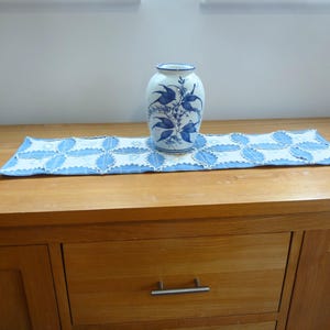 Puede incluir: Un jarrón de cerámica blanco y azul con un diseño floral se encuentra sobre un camino de mesa decorativo azul y blanco. El camino de mesa está sobre un mueble de madera con un cajón y un tirador plateado. Decoración del hogar.