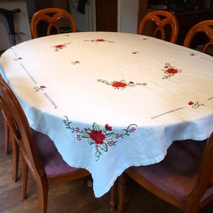 Może przedstawiać: Biały owalny obrus z haftowanymi czerwonymi różami i zielonymi liśćmi. Obrus jest położony na drewnianym stole z sześcioma krzesłami.