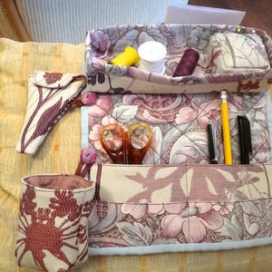 May include: A quilted fabric sewing organiser featuring a floral pattern in shades of pink, mauve, and cream. The set includes a rectangular tray with spools of thread and a pincushion, a matching cylindrical cup, a small pouch, and a flat mat with pockets holding scissors, a yellow pencil, and two black pens.