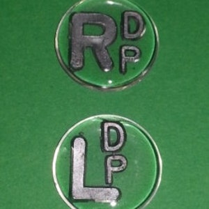 May include: Two clear, round, resin-coated tokens on a green background. The top token features the letters "RD" and "P" in black. The bottom token has the letters "LD" and "P" in black. The tokens are set against a green backdrop.
