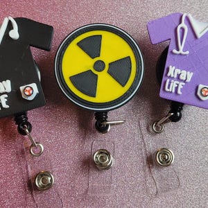 May include: Three retractable badge holders with different designs. One features a black scrub top with the text "Xray Life", another features a purple scrub top with the text "Xray Life", and the last features a yellow circle with a black radiation symbol.