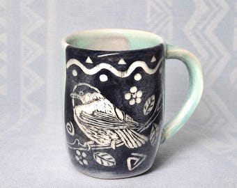 May include: A ceramic mug with a dark navy blue exterior featuring a white sgraffito illustration of a bird perched on a branch. The design includes white geometric patterns, leaves, and flowers. The interior and handle are a light mint green.