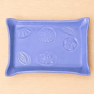May include: A rectangular periwinkle blue ceramic tray with slightly flared corners. The surface features six embossed motifs including a flower, a leaf, a fern, a seed pod, a patterned leaf, and a traditional stone lantern design.
