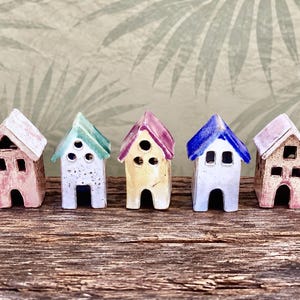 May include: A row of seven small ceramic house figurines in various colors including yellow, pink, teal, purple, blue, and white. Each miniature building features a peaked roof, a central arched doorway, and small square or circular window cutouts.