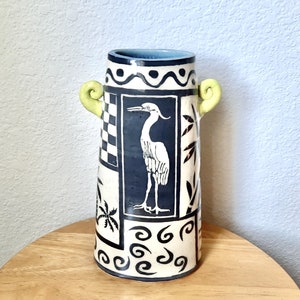 May include: A white ceramic vase with a blue and white design featuring a bird and palm trees. The vase has two green handles on the sides.