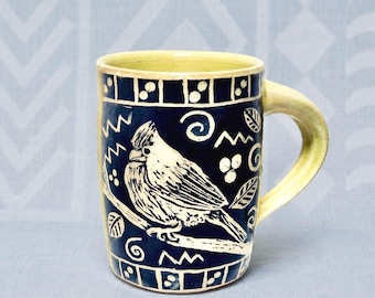 May include: A ceramic mug with a dark blue glaze featuring a white sgraffito illustration of a bird perched on a branch. The design includes decorative swirls, leaves, and geometric borders at the top and bottom. The handle and rim are light yellow.
