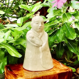 May include: A ceramic figurine of a person wearing a robe, holding a small animal in their arms. The figurine is painted in a light beige color and has a serene expression.