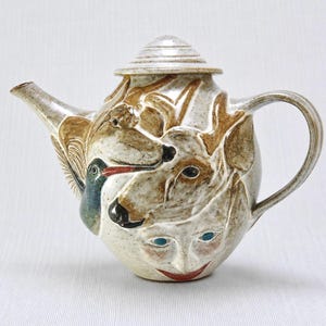 May include: A ceramic teapot with a speckled off-white glaze featuring relief carvings of a hummingbird, a deer, and a human face with blue eyes and red lips. The teapot includes a matching conical lid, a curved handle, and a tapered spout.