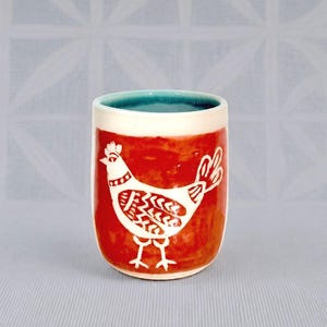May include: A cylindrical ceramic tumbler featuring a vibrant red exterior with a white graphic illustration of a chicken. The interior of the cup is glazed in a contrasting teal blue color, and the rim is finished in a clean, solid white glaze.