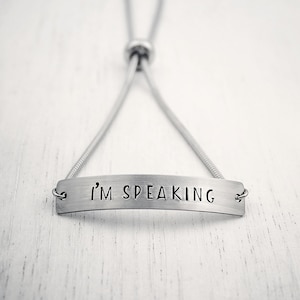 May include: A silver-toned bracelet with the phrase "I'M SPEAKING" stamped on a rectangular bar. The bracelet has a sliding clasp for adjustable sizing. The background is a white, textured surface.