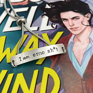 May include: A silver-toned necklace with a rectangular pendant. The pendant has the phrase "I am emo sh*t" stamped on it. The necklace is displayed on a colorful background with a book cover featuring a person.