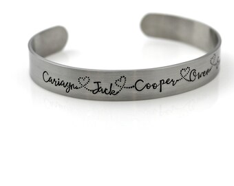 May include: A silver-colored cuff bracelet with the names Cariayn, Jack, Cooper, and Owen engraved in a cursive font. Small heart designs connect the names. The bracelet has a simple, open design.