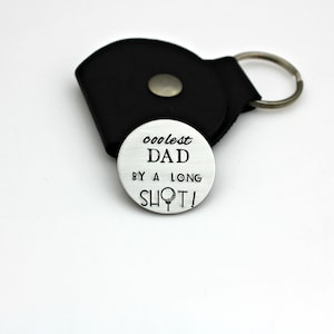 May include: A silver golf ball marker with the text "coolest DAD by a long shot!" and a golf ball and tee graphic. It is next to a black leather keychain with a metal snap and a key ring.