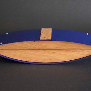 May include: A handcrafted wooden box with a curved design. The box features a vibrant blue exterior and a natural wood interior. A small wooden accent piece is secured with screws on the top. The box is approximately 30 cm long.