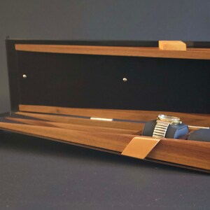 May include: Open wooden watch box with a black interior and a watch inside. The box has a slanted base and a hinged lid.