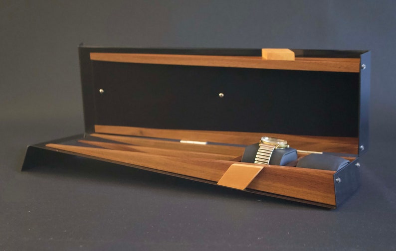 May include: Open wooden watch box with a black interior and a watch inside. The box has a slanted base and a hinged lid.