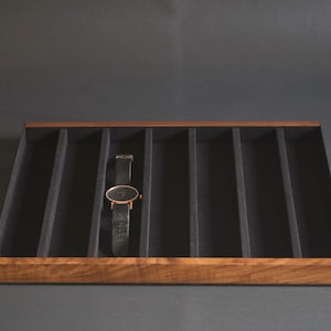May include: A wooden watch display tray with a black interior and a single watch. The tray has a dark wood frame and is divided into several compartments. The watch has a black leather band and a gold-colored watch face.