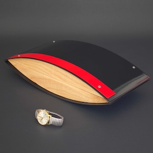 May include: A black, curved box with a red stripe and a light wood interior. A gold-toned wristwatch with a silver band is next to the box. The box has a glossy finish and is secured with small silver rivets.