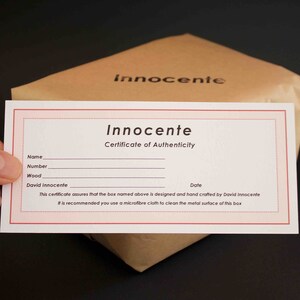 May include: A white certificate of authenticity with red borders. The certificate is for a box designed and hand-crafted by David Innocente. The text on the certificate reads: "Innocente Certificate of Authenticity Name Number Wood David Innocente Date This certificate assures that the box named above is designed and hand crafted by David Innocente It is recommended you use a microfibre cloth to clean the metal surface of this box"