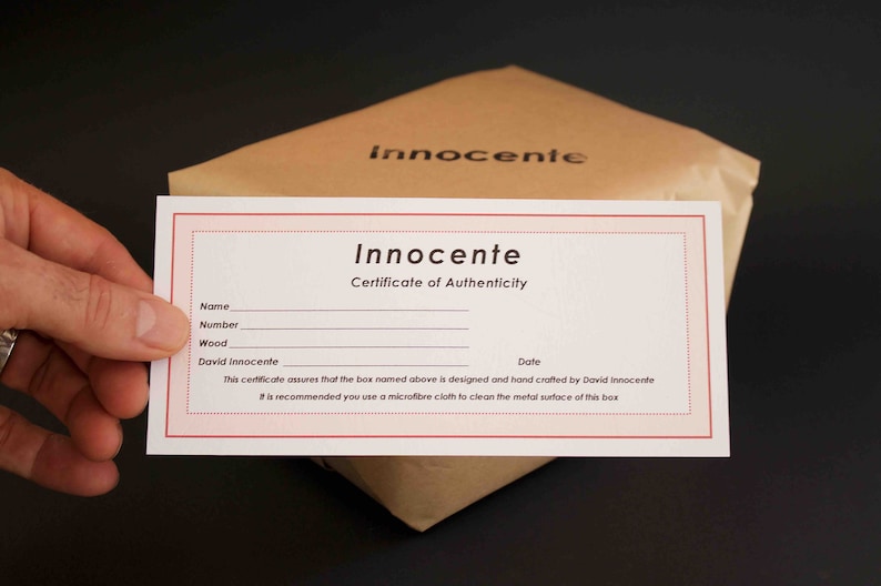 May include: A white certificate of authenticity with red borders. The certificate is for a box designed and hand-crafted by David Innocente. The text on the certificate reads: "Innocente Certificate of Authenticity Name Number Wood David Innocente Date This certificate assures that the box named above is designed and hand crafted by David Innocente It is recommended you use a microfibre cloth to clean the metal surface of this box"