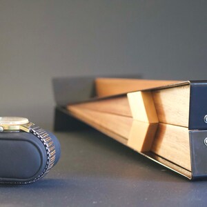 May include: A gold-toned wristwatch with a black leather band is displayed on a black watch pillow. The watch is resting on a gray surface. A wooden box with black metal accents is in the background.