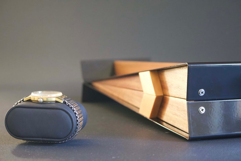 May include: A gold-toned wristwatch with a black leather band is displayed on a black watch pillow. The watch is resting on a gray surface. A wooden box with black metal accents is in the background.