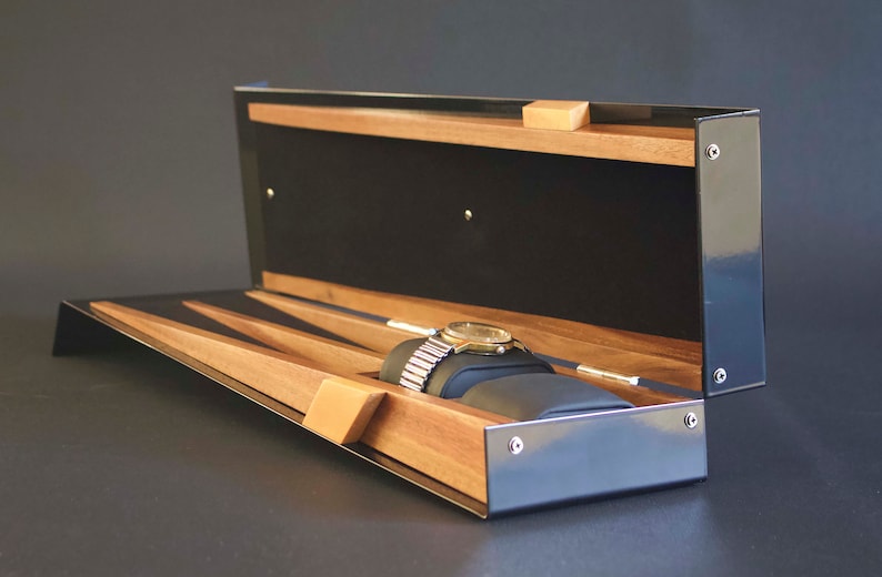 May include: A black watch box with a wooden interior and a watch resting on a cushion. The box has a hinged lid and a black exterior.