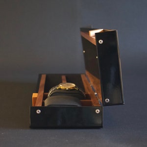 May include: A black wooden watch box with a hinged lid, open to reveal a single watch cushion and a gold watch with a black band.