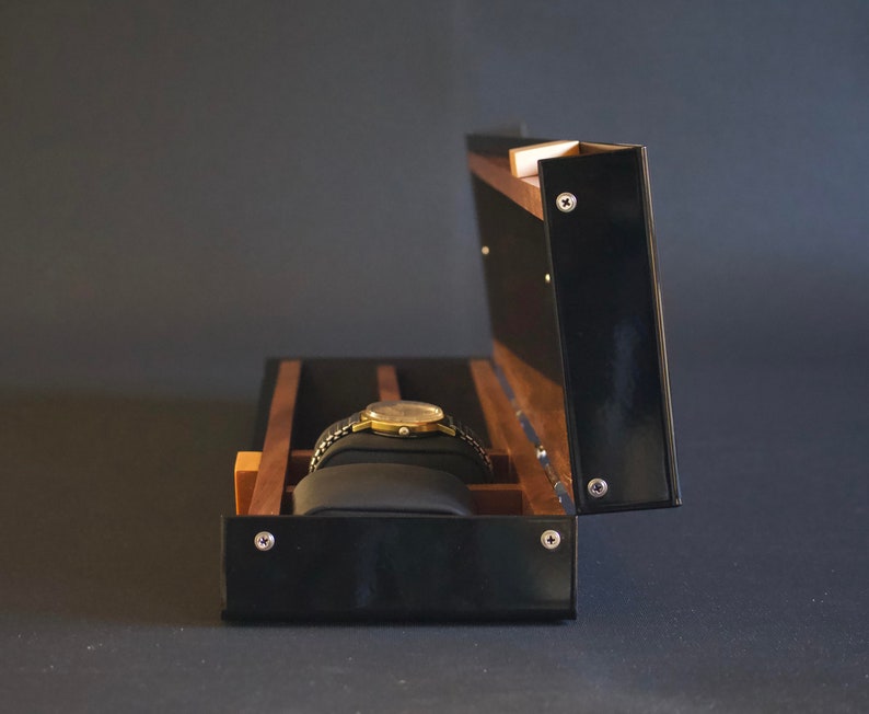 May include: A black wooden watch box with a hinged lid, open to reveal a single watch cushion and a gold watch with a black band.