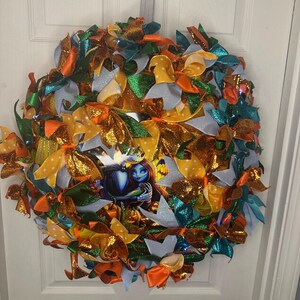 Può includere: Una ghirlanda fatta a mano con un'immagine centrale di una coppia di cartoni animati. La ghirlanda è composta da vari nastri colorati in arancione, giallo, verde e blu. Alcuni nastri hanno glitter e pois. Appesa a una porta bianca.