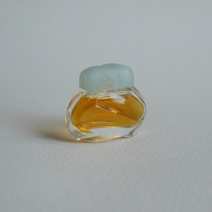 May include: A miniature perfume bottle made of clear glass, with a pale blue cap. The bottle is filled with a golden liquid. The bottle has a distinctive, rounded form, and the cap has a frosted finish. The background is a plain white surface.