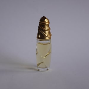 Puede incluir: Un pequeño frasco de perfume con un cuerpo de vidrio transparente y una tapa dorada. La botella tiene un diseño en espiral en el vidrio. La tapa tiene forma cónica con una banda dorada decorativa en la parte superior. El perfume en su interior es de color amarillo claro.