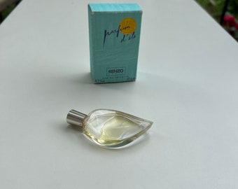 Op de afbeelding: Een heldere, blad-vormige parfumfles met een zilveren dop, naast een lichtblauwe doos. Op de doos staat "parfum d'été" en "KENZO". De fles bevat een lichtgele vloeistof. De doos is 5 ml.