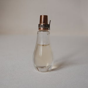 May include: A clear glass perfume bottle with a copper-colored cap and a silver band that reads "GUERLAIN". The bottle has a pear-shaped design and contains a light-colored liquid. The bottle is on a white surface.