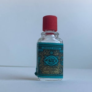 Pode incluir: Pequena garrafa de vidro transparente com tampa vermelha. A garrafa tem um rótulo turquesa e dourado com o texto "KÖLNISCH WASSER" e "No. 4711". Provavelmente é uma colônia ou perfume vintage.