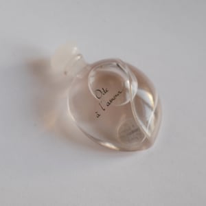 Könnte beinhalten: Eine kleine, transparente Parfümflasche mit abgerundeter Form und weißem Verschluss. Auf der Flasche steht in schwarzer Schrift "Ode à l'amour". Das Parfüm im Inneren hat eine hellrosa Farbe.