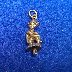 Könnte beinhalten: Ein kleiner, goldfarbener Anhänger mit einer sitzenden Feenfigur auf einem Pilz. Die Fee hat spitze Ohren und ist zusammengerollt, die Arme um die Knie gelegt. Der Anhänger hat oben eine Schlaufe zur Befestigung an einer Halskette oder einem Armband.