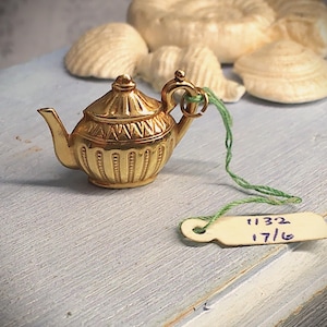 May include: A miniature gold-toned metal teapot charm featuring a pointed lid, a curved handle, and a spout. The teapot has vertical ribbed detailing and a geometric band. A green string attaches a small white tag with the handwritten text 1132 17/6.
