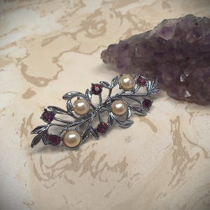 Puede incluir: Broche plateado con diseño floral, con perlas de imitación y gemas rojas. El broche tiene una apariencia delicada y ornamentada, con hojas y ramas. Un cristal púrpura en el fondo.