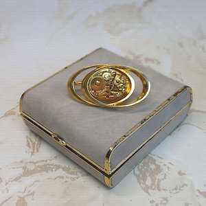 Puede incluir: Un pasador de corbata dorado con un diseño de mecanismo de reloj. El pasador tiene un marco ovalado con los intrincados engranajes y joyas de un movimiento de reloj visibles. El pasador descansa sobre una caja gris con ribete dorado.