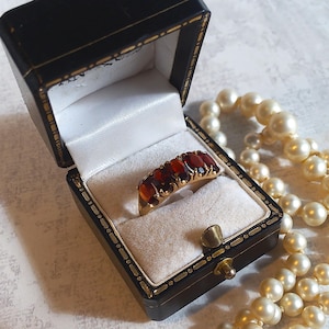 May include: A gold ring with oval-cut red gemstones displayed in a black jewelry box with a white satin interior. A strand of cream-colored pearls is draped nearby. The ring is a classic piece of jewelry.