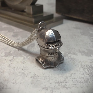 Puede incluir: Un collar con colgante plateado que representa un casco de caballero en miniatura. El casco, con su visera y remaches, está unido a una fina cadena. El colgante parece ser de metal y tiene una estética vintage o antigua. La cadena mide unos 40 cm.