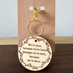 May include: A wooden keychain with a gold-colored clasp and ring. The round wooden pendant features the engraved phrase "We're here, because we're here." A decorative leaf border surrounds the text. The keychain is attached to a brown cardboard backing.