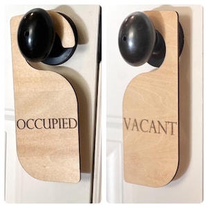 May include: A light-colored wooden door hanger with a smooth finish, featuring a circular cutout at the top to fit over a black door knob. The word OCCUPIED is engraved on one side, and the word VACANT is engraved on the other side in dark text.